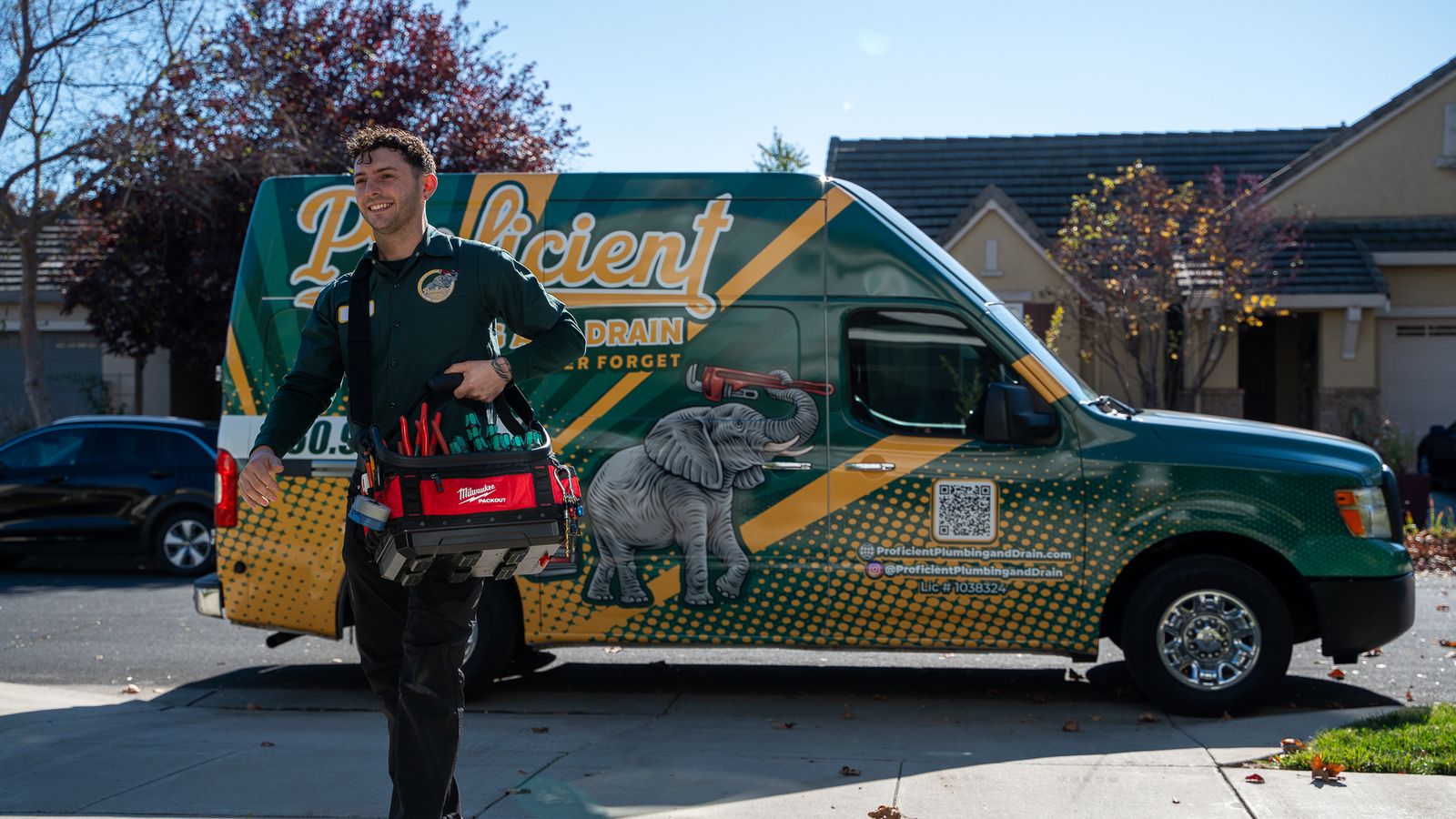 Proficient Plumbing and Drain technician walking toward the camera from a wrapped company van parked at the curb