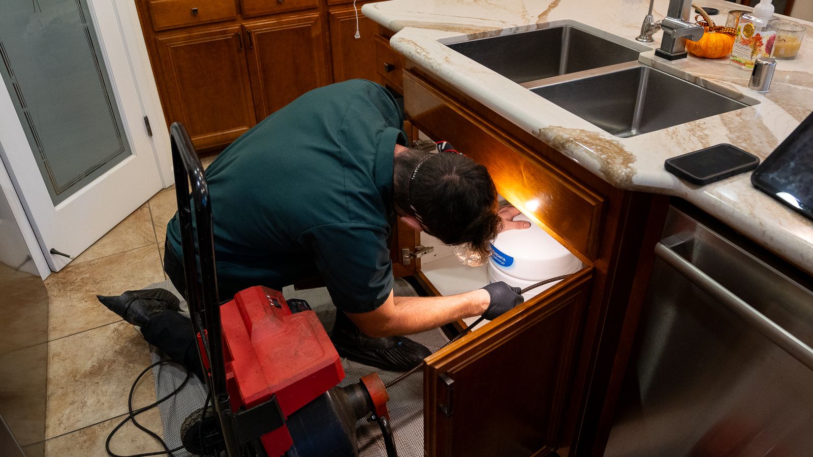 Proficient Plumbing technician working under a kitchen sink by headlamp with a drum machine running beside him