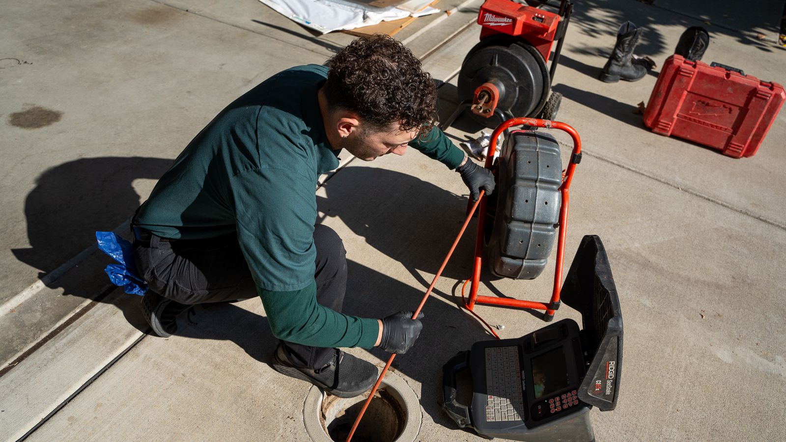 Proficient Plumbing technician feeding a sewer camera into a driveway cleanout with the monitor open on the ground beside him
