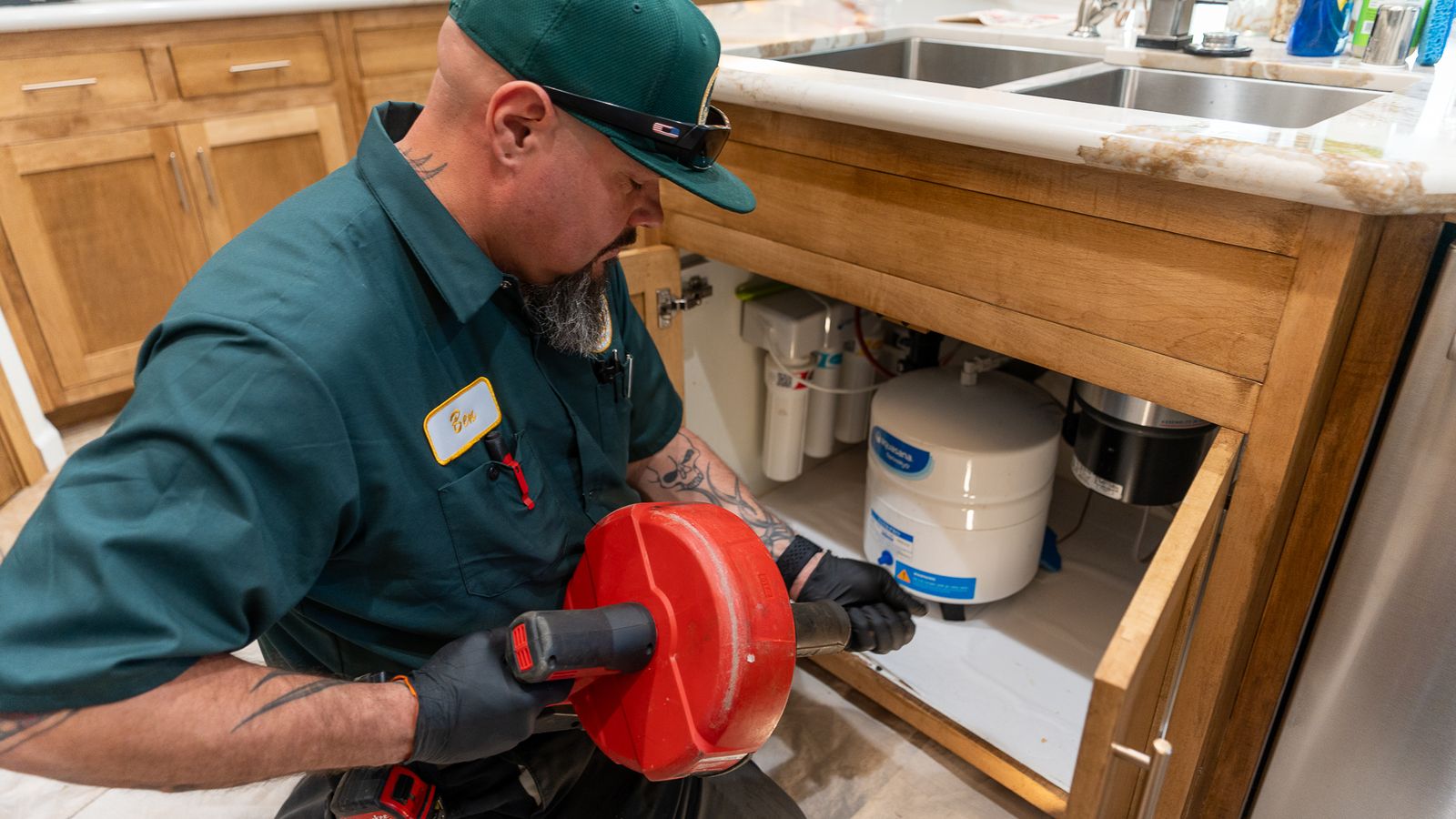 Proficient Plumbing technician kneeling at an open kitchen sink cabinet holding a drum auger