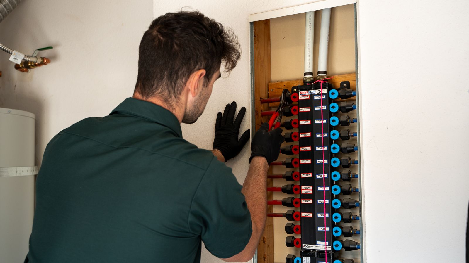 A red and blue PEX manifold mounted in a wall recess, with labelled ports and home-run lines leaving it