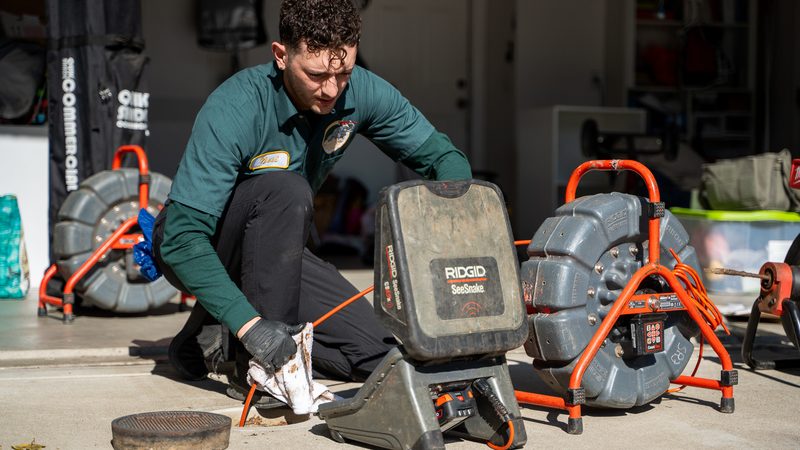 Proficient Plumbing technician setting up a sewer camera and drain machines beside an open cleanout in a residential garage