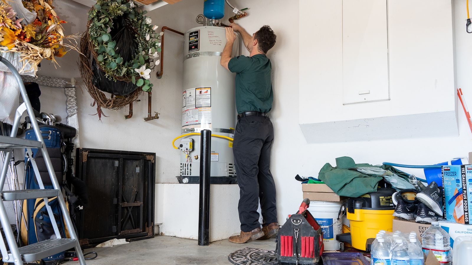 Wide view of a residential garage with a tank water heater, expansion tank, ladder and storage shelving around it