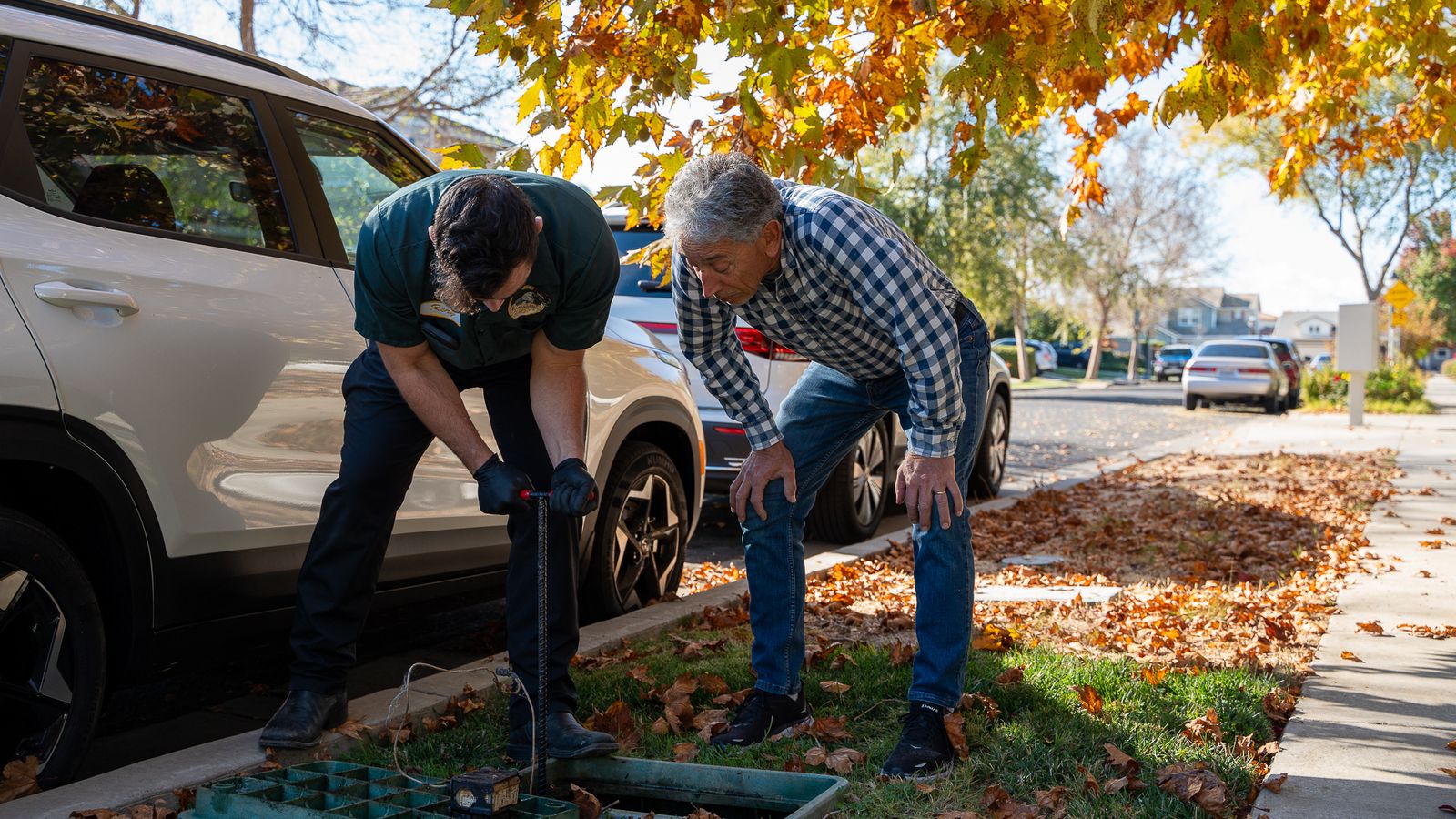 Proficient Plumbing technician working a valve key into an in-ground shutoff box while the homeowner points into the box
