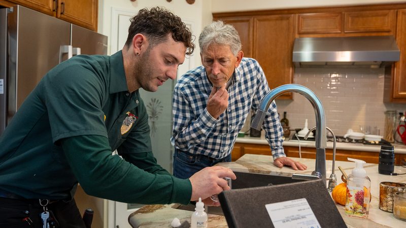 Proficient Plumbing technician running a water quality test at a kitchen sink while the homeowner looks on